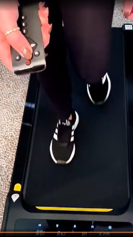 A woman walking on a walking pad while easily adjusting speed or settings with a handheld remote, without breaking stride.
