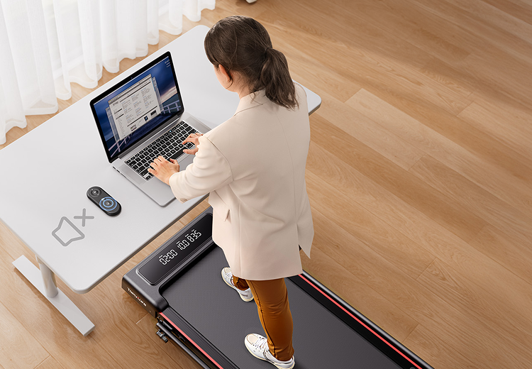 A woman works at her desk while walking on an under-desk treadmill, using the remote's mute button

